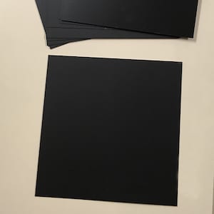 Op de afbeelding: Een stapel van tien zwarte vierkante stukjes papier, elk ongeveer 15 cm bij 15 cm, naast een liniaal.