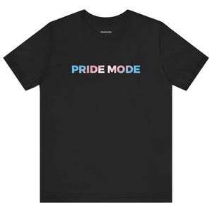 以下が含まれることがあります： 「PRIDE MODE」という文字が、青、ピンク、白のグラデーションで書かれた黒いTシャツです。