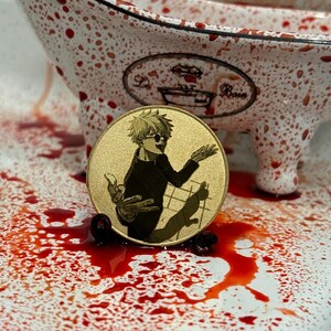 以下が含まれることがあります： 金色のコインに、金髪でサングラスをかけた漫画のキャラクターが描かれている。キャラクターは黒いシャツを着て、猫を抱いている。コインは赤い斑点のある白い表面の上に置かれている。
