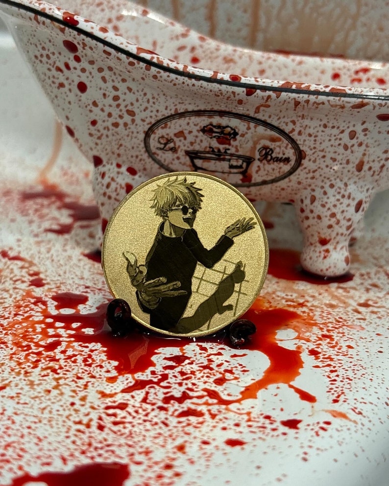 以下が含まれることがあります： 金色のコインに、金髪でサングラスをかけた漫画のキャラクターが描かれている。キャラクターは黒いシャツを着て、猫を抱いている。コインは赤い斑点のある白い表面の上に置かれている。