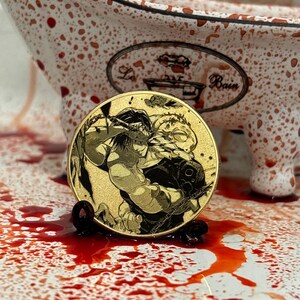 以下が含まれることがあります： 木々を背景に、剣を持った人物2人を描いた、金色のコイン。コインは赤い斑点のある白い表面の上に置かれている。