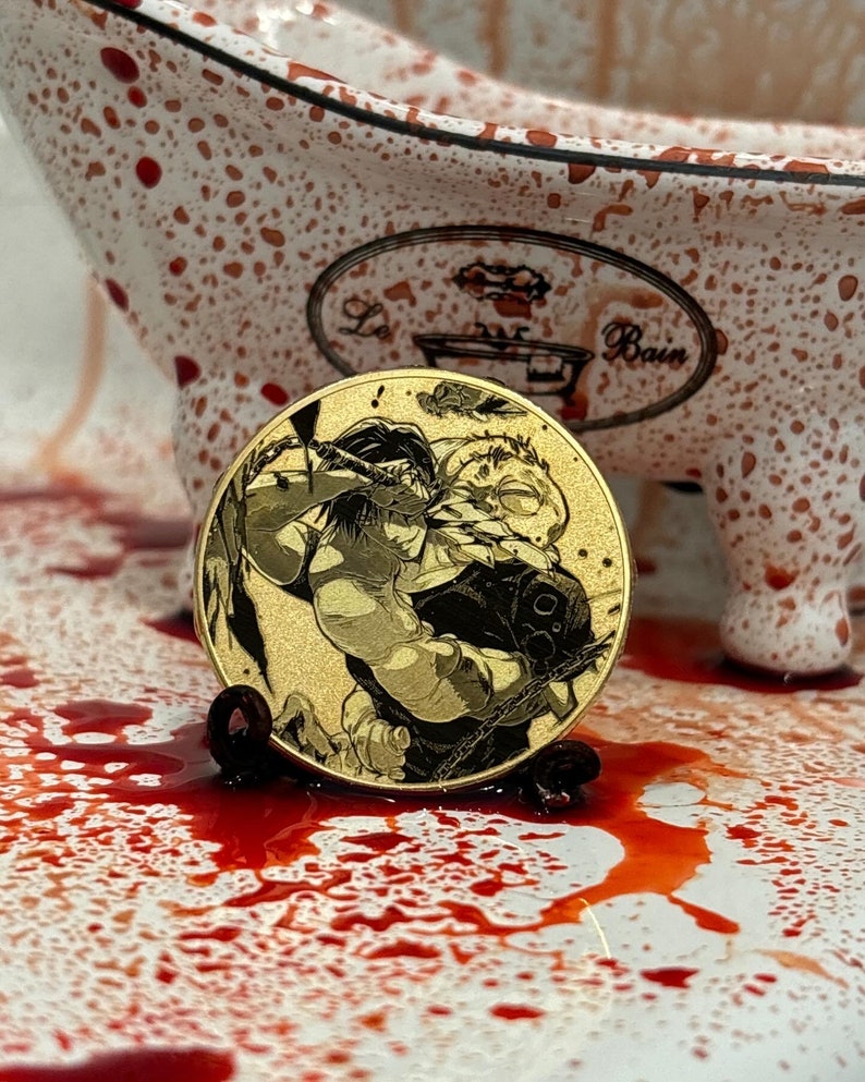 以下が含まれることがあります： 木々を背景に、剣を持った人物2人を描いた、金色のコイン。コインは赤い斑点のある白い表面の上に置かれている。