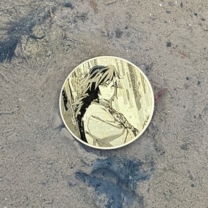 K&ouml;nnte beinhalten: Eine runde, goldfarbene M&uuml;nze mit einem Schwarzwei&szlig;bild eines Anime-Charakters mit langen schwarzen Haaren. Der Charakter tr&auml;gt ein lang&auml;rmliges Hemd und hat einen ernsten Gesichtsausdruck. Die M&uuml;nze liegt auf einer sandigen Oberfl&auml;che.