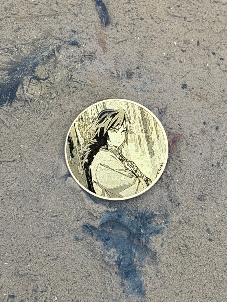 K&ouml;nnte beinhalten: Eine runde, goldfarbene M&uuml;nze mit einem Schwarzwei&szlig;bild eines Anime-Charakters mit langen schwarzen Haaren. Der Charakter tr&auml;gt ein lang&auml;rmliges Hemd und hat einen ernsten Gesichtsausdruck. Die M&uuml;nze liegt auf einer sandigen Oberfl&auml;che.