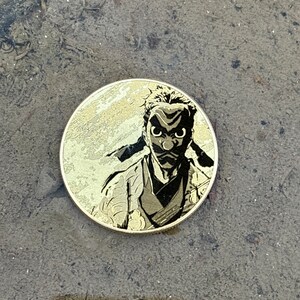 K&ouml;nnte beinhalten: Eine runde goldfarbene M&uuml;nze mit einem Schwarzwei&szlig;bild einer Person, die eine Maske und eine Robe tr&auml;gt. Die Person hat einen ernsten Gesichtsausdruck.