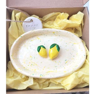Puede incluir: Un plato de cerámica hecho a mano con dos adornos en forma de limón. El plato ovalado tiene un diseño moteado amarillo y verde. Los limones son amarillos con hojas verdes. Una etiqueta con "THANK YOU" está adjunta.