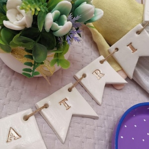 May include: A white ceramic banner spelling "ART" with gold lettering, strung on twine. The banner is in front of a small white pot of artificial flowers with green leaves and white and green flowers. A purple tray is in the foreground.