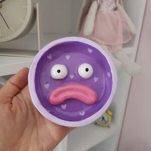 May include: A purple ceramic dish with a cartoon face design. The dish features large white eyes, a pink mouth, and small white heart accents. The dish is held in a hand, with a white shelf and a clock in the background.