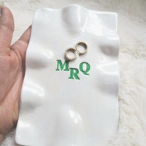 Puede incluir: Una bandeja de cerámica blanca para joyas con las letras verdes "MRQ" y dos pendientes de aro dorados con pequeñas piedras transparentes. La bandeja tiene un diseño ondulado y texturizado.