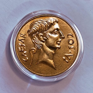 May include: A circular gold-colored coin encased in a clear plastic protective capsule. The coin features a relief profile of a person facing right, with the text CAESAR on the left and IA.702 on the right side of the central figure.