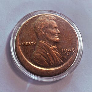 Puede incluir: Una moneda de un centavo Lincoln de 1946 de color cobre en un soporte de plástico transparente. El anverso presenta un perfil de Abraham Lincoln con la palabra "LIBERTY" arriba y la fecha "1946" abajo. La moneda está en un estuche circular.