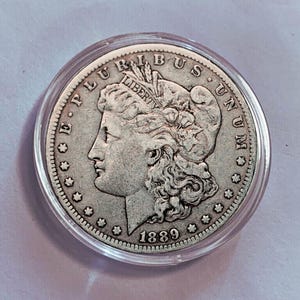 May include: A silver 1889 Morgan silver dollar coin, in a clear plastic case. The coin shows a woman's profile and the inscriptions "E PLURIBUS UNUM" and "LIBERTY". The year "1889" is displayed on the coin.
