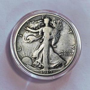 Könnte beinhalten: Eine silberne Walking Liberty Half Dollar Münze von 1919 in einer Schutzhülle aus Kunststoff. Die Münze zeigt eine Frau, die auf der Vorderseite auf die Sonne zugeht, und die Worte "IN GOD WE TRUST".