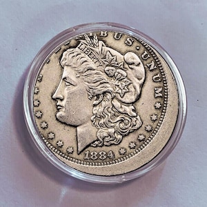 Może przedstawiać: Srebrna moneta amerykańska dolara Morgana z 1884 roku, przedstawiająca portret Wolności z koroną z warkoczy i wieńcem z liści dębu, a także napis "LIBERTY" i "E PLURIBUS UNUM".