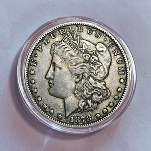 Puede incluir: Una moneda de plata de 1878, con el perfil de una mujer con cabello ondeado, rodeada por las palabras "E PLURIBUS UNUM" y "LIBERTY". La moneda está dentro de un soporte de plástico transparente.