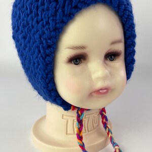 May include: A blue knitted baby hat with a long, colorful braided tie. The hat has a pointed top and covers the ears.