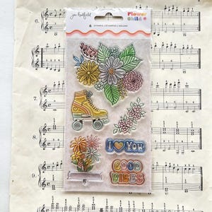 May include: Clear stamp set featuring floral designs, a roller skate, and the phrases "I Love You" and "Good Vibes." The packaging reads "Flower Child" and "6 Stamps". The stamps are on a background of vintage sheet music.
