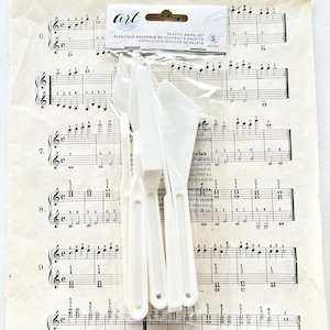 May include: A set of five white plastic palette knives in a clear package. The package has the text "art" and "PLASTIC KNIFE SET". The knives are on a sheet of vintage sheet music.