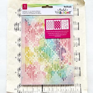 Pode incluir: Um estêncil de plástico transparente com um design aquarela colorido. O estêncil tem um padrão de linhas verticais e uma inserção retangular rosa com três padrões de diamantes. O texto "Bold + Bright" é visível.