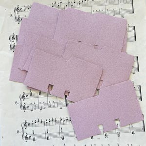 Könnte beinhalten: Eine Ansammlung hellvioletter Karteikarten mit Kerb-Tabs, die auf einem Blatt alter Noten liegen. Die Karten haben eine strukturierte Oberfläche und überlappen sich leicht. Die Noten zeigen musikalische Noten und Symbole.