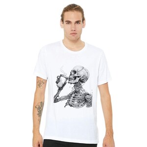 Pode incluir: Camiseta branca com um gráfico de um esqueleto bebendo café. O esqueleto é desenhado em preto e branco.