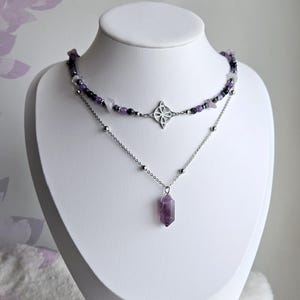 May include: Two-strand necklace set displayed on a white mannequin. The top necklace features a beaded design with purple and black stones, and a silver pendant. The bottom necklace has a silver chain with a purple crystal pendant.