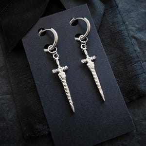 May include: A pair of silver-toned hoop earrings featuring dangling sword-shaped charms. Each earring consists of a circular hoop connected to a textured, pointed blade pendant with a cross-guard detail, displayed on a black rectangular card.