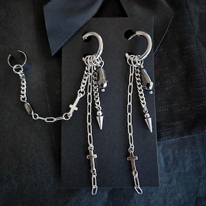 May include: Silver earrings with a dark aesthetic. The earrings feature a hoop design with dangling chains, crosses, and dark beads. One earring has an ear cuff connected by a chain.