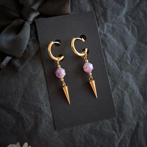 Peut inclure: Une paire de boucles d'oreilles dorées avec un design pendant. Chaque boucle d'oreille comporte une pierre précieuse facettée, violet clair et un pendentif doré en forme de cône. Les boucles d'oreilles sont présentées sur une carte noire sur fond sombre.