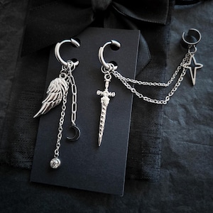 May include: Silver earrings with a gothic design. One earring has a wing charm, a crescent moon, and a dangling crystal. The other earring features a sword charm. A third piece has a star charm connected by a chain.
