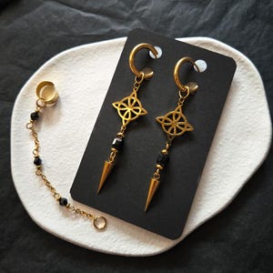 May include: A pair of gold-toned hoop earrings featuring a central knot charm, a black bead, and a gold-toned spike pendant. A separate gold-toned ear cuff is connected by a delicate chain adorned with small black beads to the earring set.