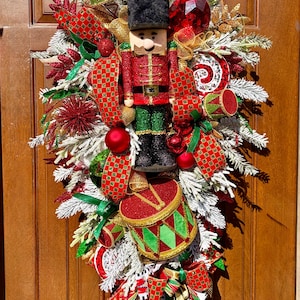 May include: A festive Christmas door decoration featuring a nutcracker soldier. The wreath includes red, green, and gold ribbons, ornaments, and faux snow-covered branches. A large red drum and a red gem add to the holiday cheer.