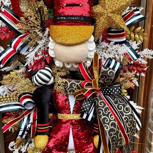 May include: A festive Christmas wreath featuring a nutcracker design. The wreath is adorned with red sequins, gold accents, and various ribbons in black, white, and red. A gold star and decorative elements complete the holiday theme.