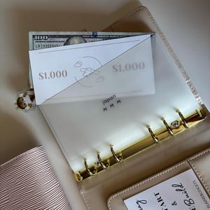 May include: A beige planner with gold-colored rings, holding a $100 bill and two cards labeled "$1,000". A clear plastic divider is inside the planner. The text "japan" and a small illustration are visible on the divider. A small charm is attached to the planner.