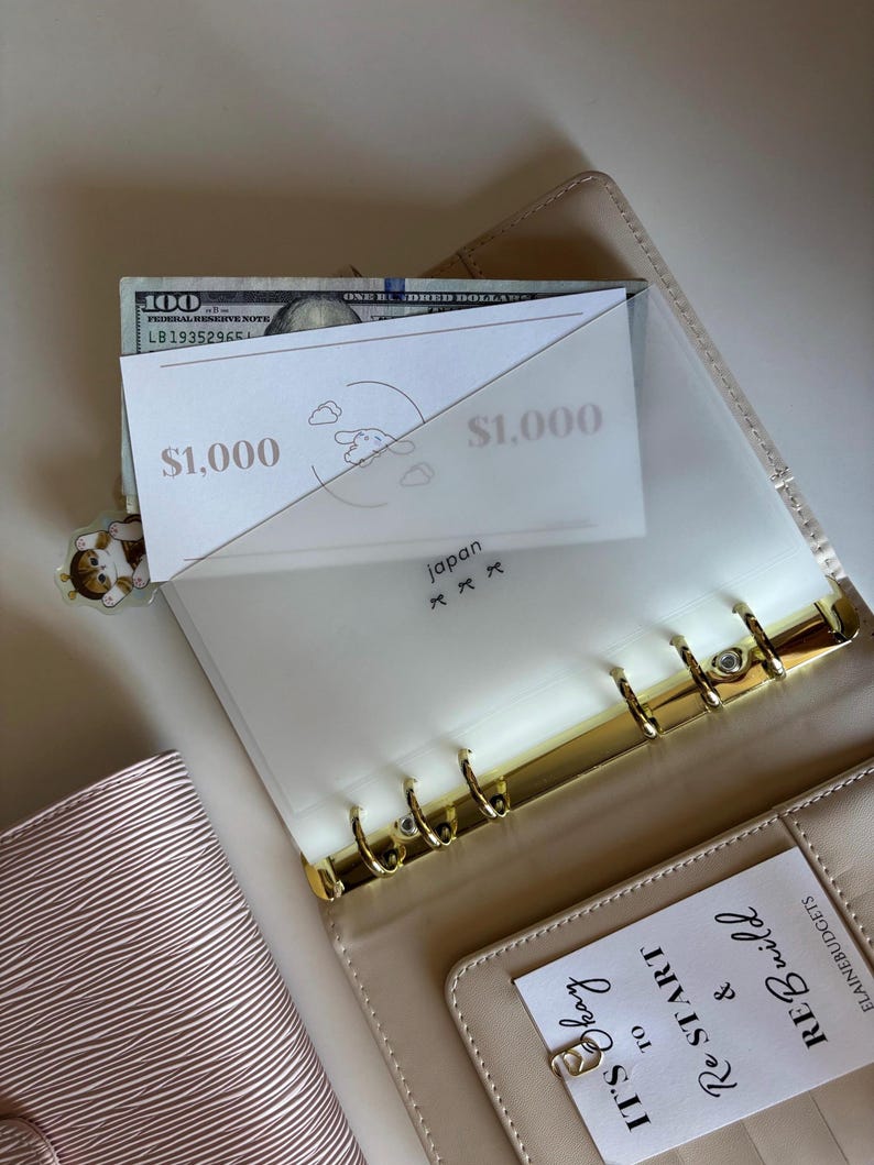 May include: A beige planner with gold-colored rings, holding a $100 bill and two cards labeled "$1,000". A clear plastic divider is inside the planner. The text "japan" and a small illustration are visible on the divider. A small charm is attached to the planner.