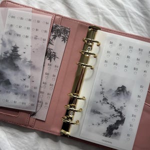May include: A pink six-ring binder containing translucent vellum inserts with black ink illustrations of pagodas, mountains, and cherry blossoms. Each insert features a grid of circles paired with dollar amounts for financial tracking.