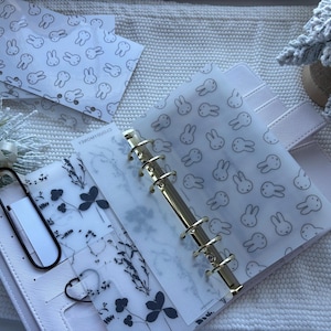 May include: A white planner with gold hardware, open to show decorative inserts. The inserts include a bunny pattern and floral designs. Several matching bunny-patterned envelopes are visible.