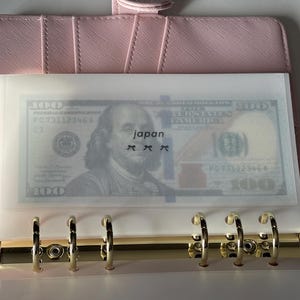May include: A pink binder with gold rings holds a clear plastic sleeve containing a US $100 bill. The word "japan" is printed on the sleeve. The binder is open, revealing the rings and a portion of the interior.