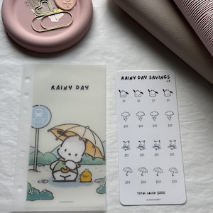 May include: A clear plastic binder insert with the text "RAINY DAY" and a cartoon character under an umbrella. A savings tracker card with "RAINY DAY SAVINGS" and illustrations of rain, characters, and umbrellas. A pink dish with decorative items.