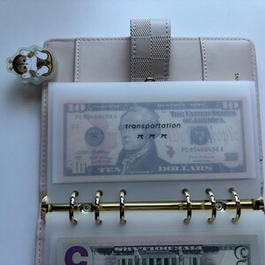 May include: A light beige planner with gold-colored rings, holding a clear plastic sleeve with a ten-dollar bill and the word "transportation". A cat-shaped sticker is attached to the planner.