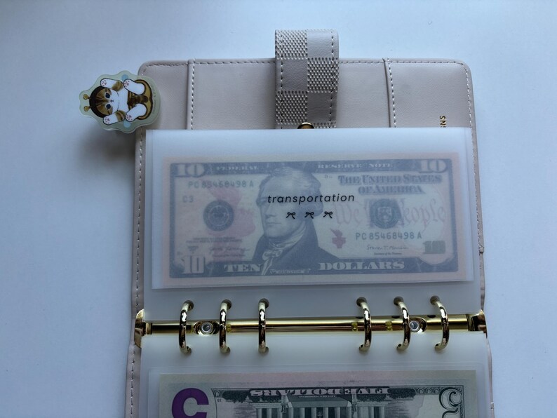 May include: A light beige planner with gold-colored rings, holding a clear plastic sleeve with a ten-dollar bill and the word "transportation". A cat-shaped sticker is attached to the planner.