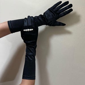 May include: Black satin opera gloves with a pearl ring. The gloves are long, reaching the upper arm, and have a smooth, elegant texture. The ring features a row of white pearls. The gloves are displayed against a neutral background.
