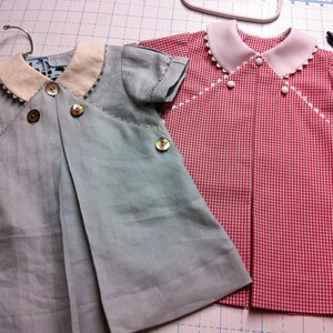 May include: Two baby dresses, one light blue with white trim and buttons, and one red and white gingham with white trim and buttons.