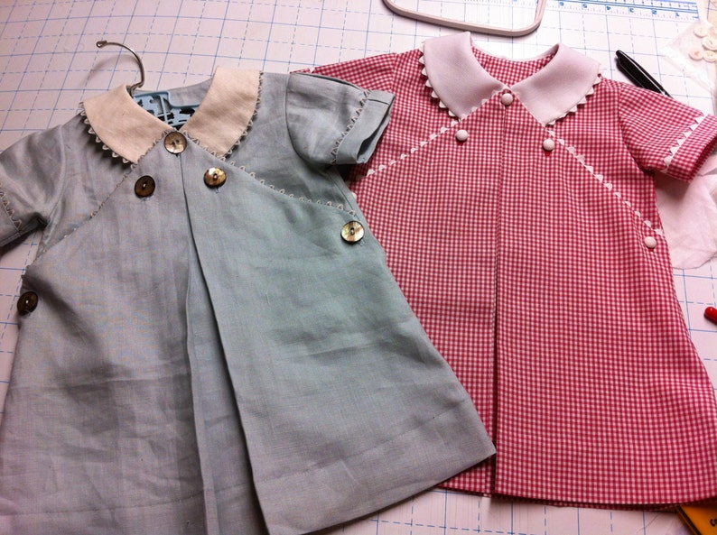 May include: Two baby dresses, one light blue with white trim and buttons, and one red and white gingham with white trim and buttons.