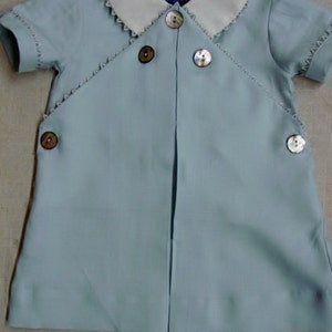 May include: A light blue baby outfit with a white collar and pearl buttons. The outfit has a scalloped trim around the collar and sleeves.
