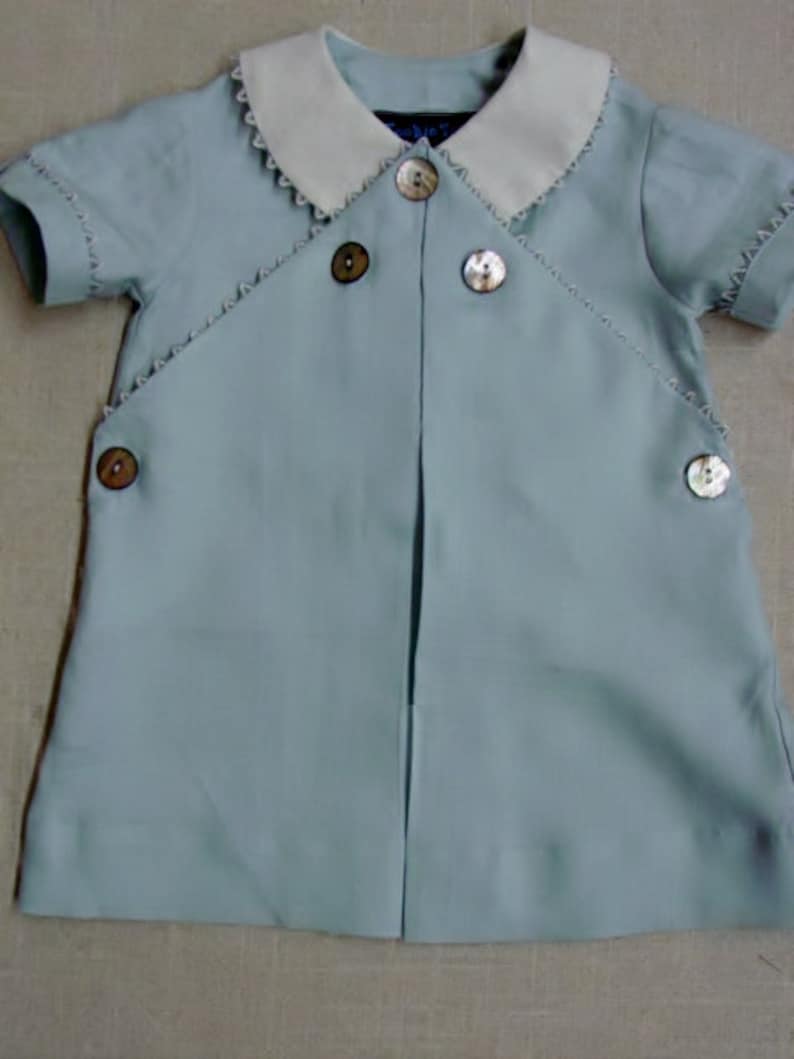 May include: A light blue baby outfit with a white collar and pearl buttons. The outfit has a scalloped trim around the collar and sleeves.