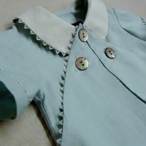 May include: A light blue baby outfit with a white collar and pearl-like buttons. The outfit has a scalloped edge and is made of a soft, lightweight fabric.