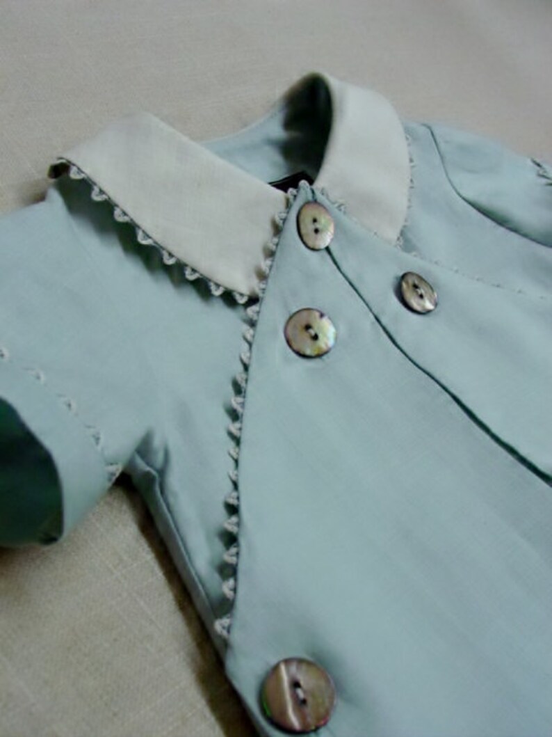 May include: A light blue baby outfit with a white collar and pearl-like buttons. The outfit has a scalloped edge and is made of a soft, lightweight fabric.