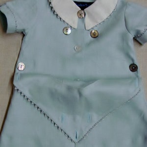May include: A light blue baby outfit with a white collar and scalloped edges. The outfit has a button closure and a decorative flap with scalloped edges.