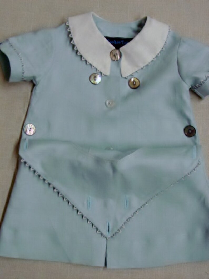 May include: A light blue baby outfit with a white collar and scalloped edges. The outfit has a button closure and a decorative flap with scalloped edges.